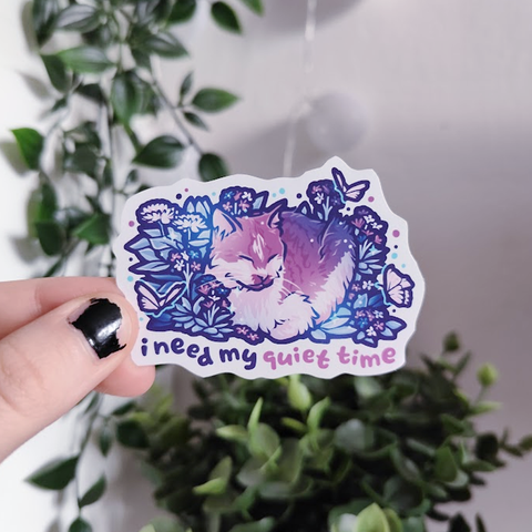 hand holding up a sticker of a kitty loafing in between flowers and butterflies, titled "i need my quiet time"