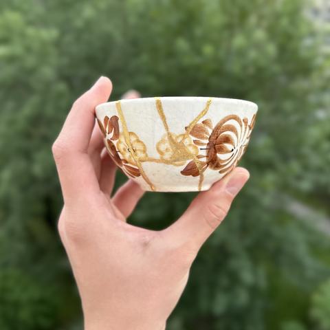 Hand holding a small bowl; its colour is ivory-white, with hand-painted brown and dark yellow flowers of various shapes. Bold gold lines keep the pieces together. Side view.