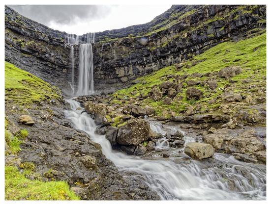 A stunning waterfall cascades down a rocky cliff, surrounded by lush green grass and boulders. The smooth movement of water flows over stones, creating a serene and picturesque natural scene. Clouds hover above, adding to the dramatic atmosphere.