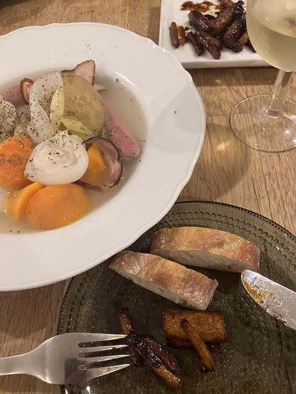 A plate of stew with vegetables including carrots, potatoes, and onions, accompanied by slices of bread and a fork. Nearby, there's a small plate with glazed vegetables and a glass of white wine. The setting features a wooden table.