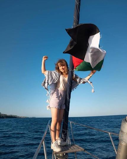 Greta Thumberg at the bow of the Madleen at sea with her right fist held high, and hel left hand holding the flag of Palestine. She's wearing shorts and a t-shirt and a kefifyeh.