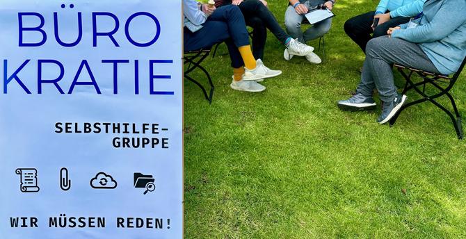 Ein Banner steht auf einer Wiese. Auf dem Banner steht in großen blauen Buchstaben: „BÜROKRATIE“. Darunter in schwarzer Schrift: „Selbsthilfegruppe – Wir müssen reden!“ Vier Symbole sind darunter abgebildet: eine Papierrolle, eine Büroklammer, eine Cloud und ein Ordner mit Lupe. Rechts vom Banner sitzen mehrere Personen auf Gartenstühlen in einem lockeren Kreis auf dem Rasen, nur ihre Beine und ein paar Unterarme sind zu sehen. Die Szene wirkt informell und einladend – wie ein Gespräch im Freien bei einer Veranstaltung.