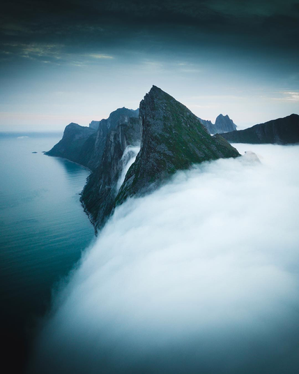 Vue aérienne spectaculaire de la montagne Segla à Senja, en Norvège, émergeant au-dessus d’une mer de nuages, avec des falaises abruptes plongeant vers l’océan sous un ciel bleu-gris.