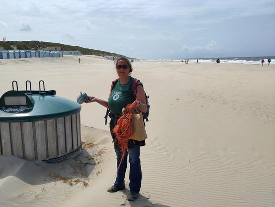 Afvalcontainer op strand waar ik rechts naast sta met opgeraaptafval. Aluminiumfolieballon in de ene hand, papieren tasje vol afval in de andere hand en een stuk oranje visnet. Op de achtergrond links duinen en strandhuisjes en rechts de zee.