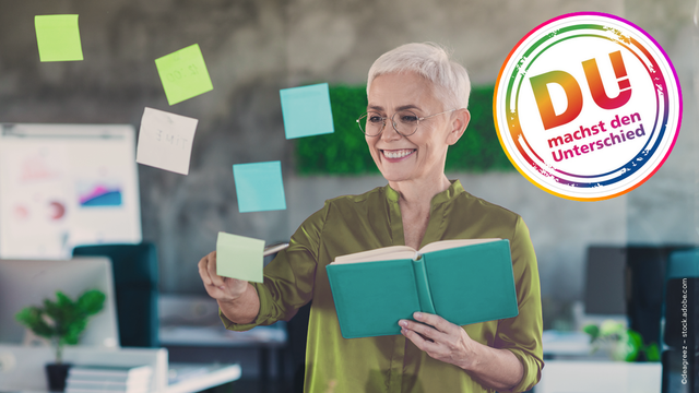 An older woman with short white hair and glasses, wearing a green blouse, is standing in a modern office space. She is smiling while holding an open green notebook and placing colorful sticky notes (blue, green, and white) on a transparent glass board. Behind her, there are computer screens, a plant, and a blurred office background. In the upper-right corner, there is a vibrant circular logo with a rainbow-colored border containing the German text "DU machst den Unterschied" ("YOU make the difference"). The scene conveys a positive, engaged, and empowering work atmosphere.