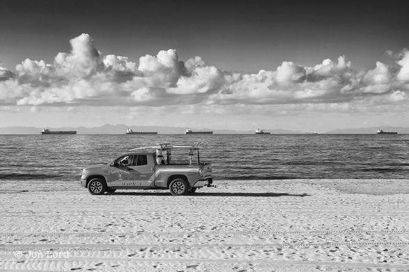 This Is A Black And White Landscape / Seascape Photo In Landscape Format. In The Foreground Rising Upwards One Third In The Frame Is A Smooth And Level Beach. To The Left Of Centre Is A Lifeguard Pickup Truck With A Surfboard On Top And Moving From Right To Left. Beyond Is The Ocean Rising Upwards To The Half Way Point Where The Sea Meets A Hazy Undulating Ridge Of Terrain, Perhaps Thirty Of Forty km Away. Before This Terrain Resting On The Horizon, About Five Or Ten km Away Is A Line Of Five Anchored Cargo Ships, Probably Crude Oil Tankers, All Viewed In Profile With Their Bows Pointing To The Right. Half Way Along The Right Side Is The Speck Of A Further Cargo Ship Probably Underway And Moving Away From The Camera. Above Is A Broken Layer Of Stratocumulus Cloud, With Clear Sky Above. 
The Location: Huntington Beach, California. The Distant Terrain Is Santa Catalina Island. The Body Of Water Is The San Pedro Channel (Pacific Ocean) (2015).