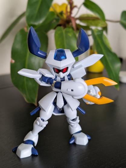A completed white and blue Rokusho Medabot model kit, standing on a dark surface in front of a green plant. Rokusho has large blue horns, red eyes, and is holding a yellow sword-like weapon in one hand.