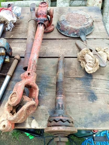 Dirty table full of stuff. The stuff includes a large red PTO shaft with two universal joints and splined ends. There's also a bent drive shaft with a clutch on it, and on the left half of a hydraulic cylinder with a rag wrapped around the end. A small piston without cylinder as well. A very crusty steel pan at the top and some kind of turning handle for a keyed shaft.