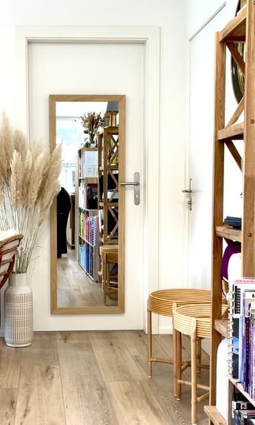 Ein Spiegel hängt in einem Raum mit Holzfußboden und Bücherregalen rechts im Vordergrubd. Links vom  Spiegel eine Vase mit Pampasgrass.