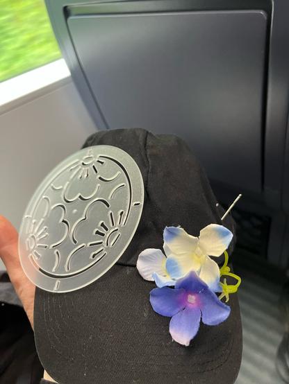 Die schwarze Schirmmütze von vorn. Es liegen eine Sashiko Schablone und mehrere violett-blau-beige Plastikblumen auf ihr.