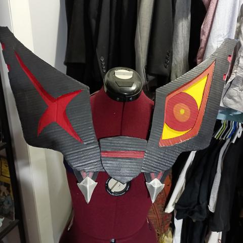 A cardboard chest piece on a seamstress mannequin. Each side has a giant shoulder piece, the left has a red x on it and the right has a red, orange, and yellow eye