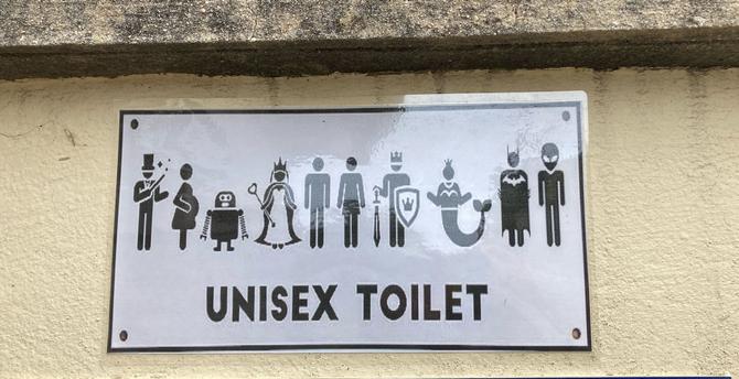 Sign for a unisex public toilet with a variety of symbols including mermaid, alien, superhero as well as the standard ones for man and woman above the words ‘UNISEX TOILET’.