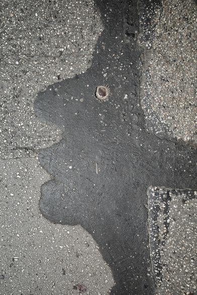 The image shows a close-up of an asphalt surface with a patch of darker, smoother tar forming an irregular shape. Embedded near the center is a rusted bottle cap. The surrounding area consists of rough, speckled concrete with small stones and debris.