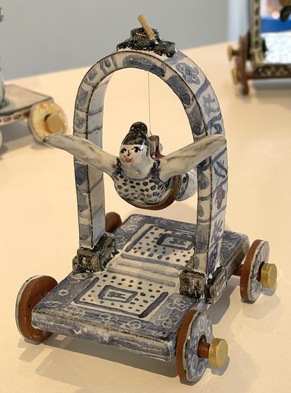 Clay sculpture of a woman in swimming costume suspended from an arch over a small wheeled cart.