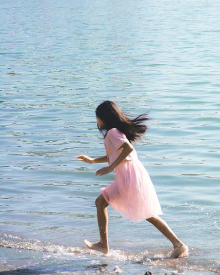 Girl in a pink dress runs along the shoreline of a large body of water. Her hair flows backward as she moves, and small splashes form around her feet. The water is calm with gentle ripples reflecting the sunlight.