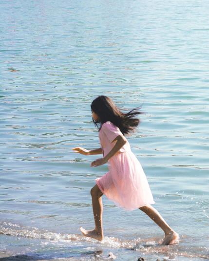 Girl in a pink dress runs along the shoreline of a large body of water. Her hair flows backward as she moves, and small splashes form around her feet. The water is calm with gentle ripples reflecting the sunlight.