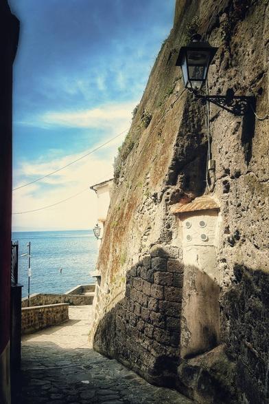 A narrow, sunlit stone path descends alongside a tall, weathered stone wall on the right, leading towards the bright blue sea visible in the distance. A classic black street lamp is affixed to the wall, casting a shadow. On the left, another stone structure frames the view. The sky above is a clear blue with scattered white clouds. The overall mood is bright, warm, and inviting.
