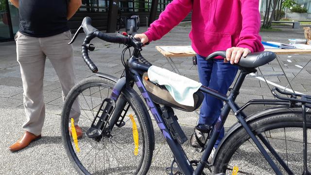 Vélo Gravel TREK dans les mains de sa propriétaire venu à l'atelier pour apprendre à démonter ses roues et plaquettes de freins. Un homme se tient proche légèrement à l'arrière-plan.