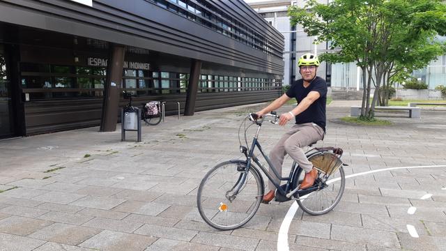 Un homme essaie un vélo de ville type hollandais sur la place de l'Europe d'Hérouville. On voit un bout de marquage au sol de la piste vélo-école à droite.