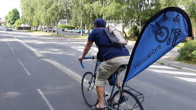 Cycliste sur un vélo équipé d'un mat porte-comm et d'un  drapeau fixé à ~1.5m de haut, sur un trottoir cyclable entre ombres des arbres et soleil.