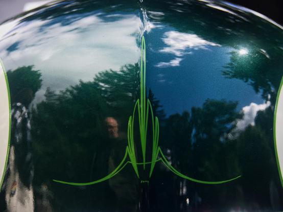 Close-up of a shiny, dark green car hood reflecting trees, sky, and clouds. In the center of the hood is a symmetrical pinstripe design in neon green.