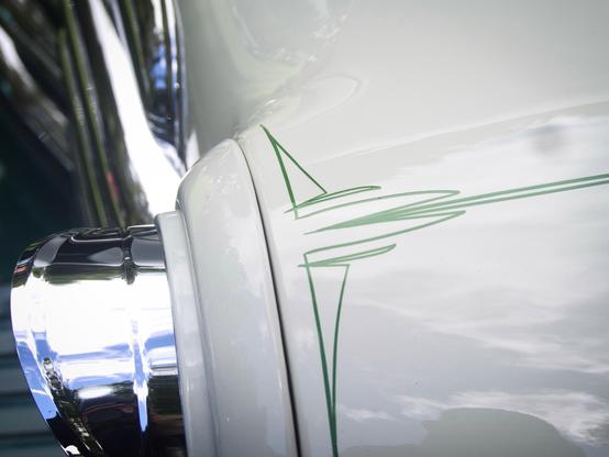 Close-up of a classic car’s white painted body with green pinstripe detailing, and a shiny chrome bumper reflecting light and surroundings.