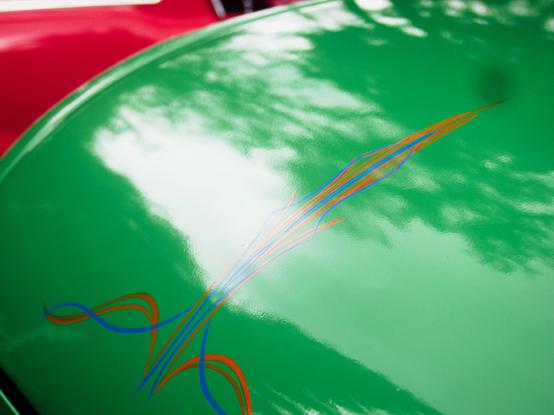 Shiny green car hood with intricate orange and blue pinstriping designs. The hood reflects the sky and trees above. A red car is partially visible in the background.