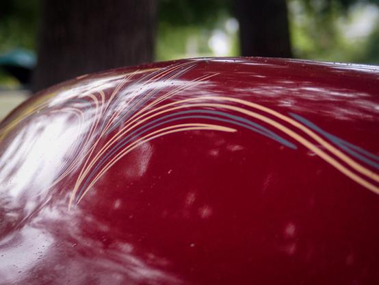Close-up of a shiny red car surface featuring decorative yellow and white pinstripes; background shows blurred trees and greenery.