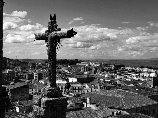 Una cruz de piedra erosionada por el tiempo, con una textura rugosa y musgo, se alza sobre una vista panorámica en blanco y negro de una ciudad. Los tejados de las casas, de un estilo arquitectónico similar, se extienden hacia el horizonte bajo un cielo nublado. En la distancia, se vislumbra una llanura extensa. La imagen transmite una sensación de antigüedad y serenidad.