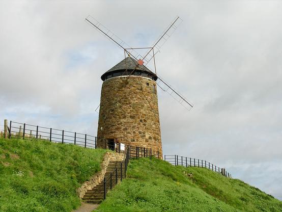 St Monans Windmill. The image shows an embankment covered in low vegetation from near the camera on the left to further away on the right. A set of stone steps leads up it left of centre and at the top is a stone windmill in the centre of the frame. There are railings along the top of the embankment. The scene is in sunlight.