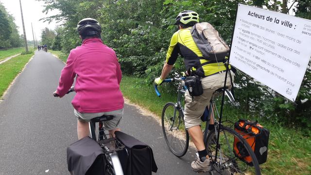 Cyclistes femme et homme sur la voie verte de Caen à Ouistreham. Un grand panneau fixé à un mat à l'arrière d'un des vélos dit : Oyé 
"Acteurs de la ville (Contrat de ville)
• Meilleure santé des jeunes : avec moins de    voitures
• Plus d'égalité femmes / hommes : aidée par des itinéraires vélo / PMR sécurisés
• Stages et emploi sont rarement de l'autre côté  de la rue : insertion
• Rouler en VAE : *si* abri vélo au domicile
• Meilleure image, regard et attractivité de l'habitat pour  les (jeunes) ménages"