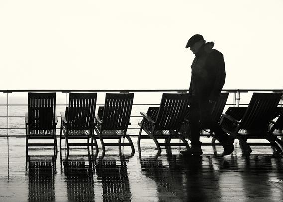 A person in a hat and coat walks on a ship's deck. The deck is wet, reflecting rows of empty wooden deck chairs. The background shows the ocean and a cloudy sky.