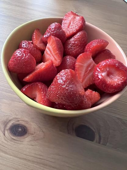 Eine Schale mit frischen, halbierten Erdbeeren steht auf einem Holz-Esstisch 😋