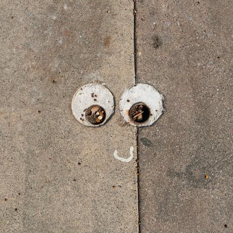 A section of sidewalk where two circular metal pipe fittings filled with debris resemble cartoonish eyes. Below them, a small curved white straw wrapper mimics a smiling mouth, creating a face-like illusion in the pavement.