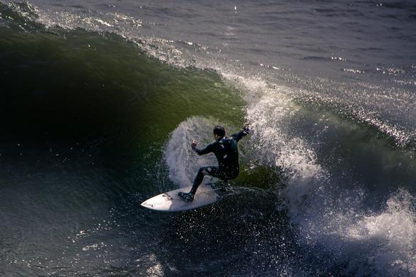 A surfer in a black wetsuit carves along the face of a large green wave in Santa Cruz, California, leaving a spray of water behind.