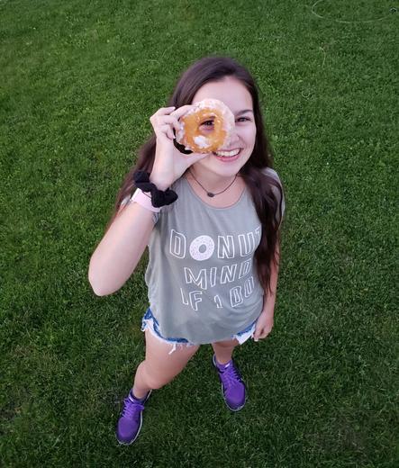 Emily holding donut up to her eye and looking through the donut hole. Picture taken from above looking down on her. She is wearing a grey tee shirt that says "Donut Mind If I Do", short denim shorts and purple sneakers.