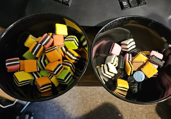 two bowls of liquorice allsorts, they're both black bowls, on the left all of them are of the type the made of layers of soft candy and liquorice with two layers of licorice sandwiched between three layers of soft candy, these are in four colours with the outer being orange pink yellow and green candy all with a central white candy. the bowl on the right contains a broader variety of all sorts with a couple that are made up of the same style of layers as the one on the left but with either all white candy or a yellow central candy, there are also some that have two layers of candy sandwiching one layer of liquorice, these are in pink orange or brown, there is also a shaped basic licor stick, electric stick with a central white candy, and some little black licorice figurine, and finally a small lozenge covered in tiny blue hundreds and thousands

the ones in the left bowl look generally fatter and fuller, the ones in the right bowl kind of look like they've been sat on by Jabba the Hutt