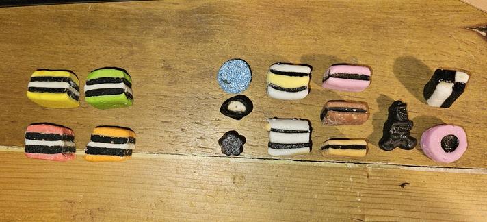 two groupings of licorice allsorts, on the left are four that are made up of layers of candy and liquorice with two layers of candy around two layers of liquorice, they are in four colors with the outer candies being yellow, green, pink, and orange all with a central white candy 

on the right is a larger range of slightly more squashed looking allsorts, these consist of a round one covered in tiny blue hundreds and thousands, a tubular one with a white candy center, a flower shaped short tubular plain liquorice, two three candy layer ones with a yellow center in one and all white on the other, three that are two layers of candy around one layer of licorice with one being pink one being brown and one being orange, a strange little licorice figurine with a hat on, a round pink candy with a round licorice centre, and a four-square square ended allsort with two white squares opposite two liquorice squares