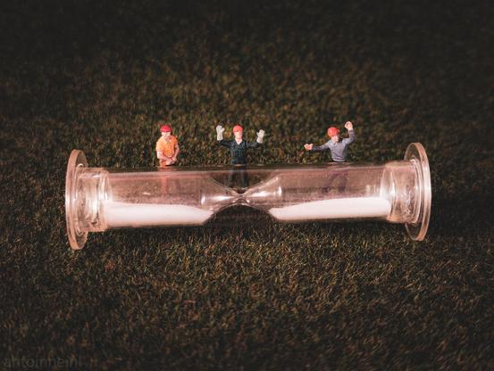 A miniature hourglass lying horizontally on a green, textured surface. There are three small miniature construction workers  positioned around the hourglass. The hourglass itself is clear glass, with white sand inside. The sand is settled in both bulbs since it's lying flat.