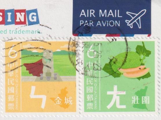Stamps of a postcard from Taiwan. The left is orange and shows something that looks liek a dog or a dragon statue wearing a red cape standing in front of grey cups with green mountains in the back. The second one is green and shows a green fruit. An Air Mail sticker is above the stamps.