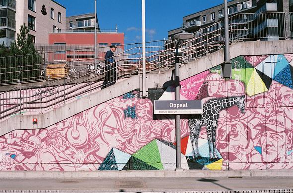 Norsk: Fargefoto av gatekunst ved trappene på Oppsal t-banestasjon. Betongveggane ved plattforma er måla med portrett av teikneserie-aktige karakterar i rosa: superheltar, hundar, ei jente med hovudtelefonar og fleire andre. Innimellom er også fargerike geometriske mønster – og ein sjiraff i svartkvitt.

Ovanfor all gatekunsten går ein eldre mann opp trappene frå plattforma, og fleire moderne bustadblokker synest i bakgrunnen. Det er sol og blå himmel, og mannen har både solbriller og skuggelue.

English: Colour photo of street art by the stairs at Oppsal metro station in Oslo. The concrete walls by the platform are painted with portraits of comic-styled characters in pink: super heroes, dogs, a girl with headphones and more. In between the characters are colourful geometric shapes – and a black-and-white giraffe.

Above all the street art, an older man walks up the stairs from the platform, with several modern apartment blocks in the background. It's sunny and the sky is blue, and the man is wearing both sunglasses and a cap.
