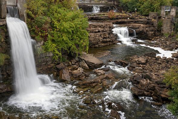 A series of waterfalls, some small, some larger, cascade over various rock shelves along the Mississippi River in Ontario, Canada.  The larger foreground waterfall has a large bush providing a green covering to the surrounding rocks.