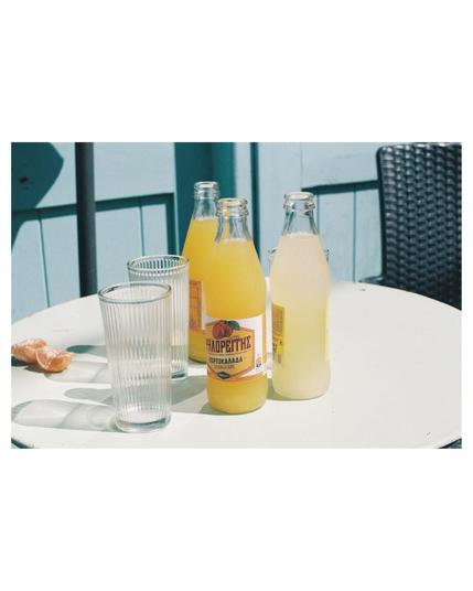 Three full sodas, two orange and one lemon, in glass bottles are standing on a round light green table with extra empty glasses and some peeled mandarins. Light blue painted wooden background and a small part of the back rest of black chair in the right upper corner. The photo is bright and sunny, through the glasses on the table are some reflections on the table surface visible