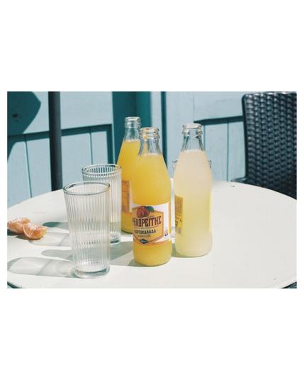 Three full sodas, two orange and one lemon, in glass bottles are standing on a round light green table with extra empty glasses and some peeled mandarins. Light blue painted wooden background and a small part of the back rest of black chair in the right upper corner. The photo is bright and sunny, through the glasses on the table are some reflections on the table surface visible