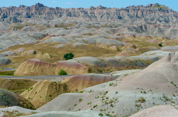a landscape loaded with geological features is shown in this photograph. the land takes up most space, with a tiny sliver of blue sky at the top above a jagged row of rocks, made of materials like  sandstones, siltstones, mudstones, claystones, limestones, volcanic ash, and shale. in the forground, rolling mounds reveal layers of various colors including green and orange