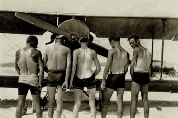 5 men pose in font of a biplane. They have removed their shirt to show their tan lines.