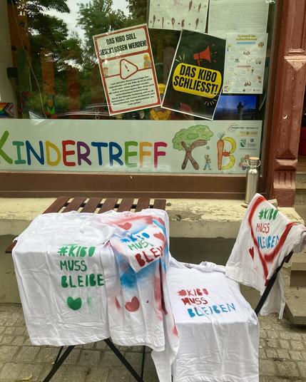 Vor dem Schaufenster des Kindertreff KiBos steht ein Gartenstuhl und ein Tisch, behängt mit weißen Ti-Shirt auf die Sprüche in verschiedenen Farben gesprüht wurden: #KIBO MUSS BLEIBEN