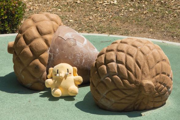 Photo of the Creature Mastodon stuffed toy next to concrete sculptures of acorns the size of Golden Retrievers at a park during one bright afternoon. The Creature is facing the camera, its back against an acorn pointed towards image-right, and image-right of that is the second acorn pointed towards the background, its cupule facing the camera. There are signs of old white graffiti washed off the sun-cracked brown stain on the concrete nuts.