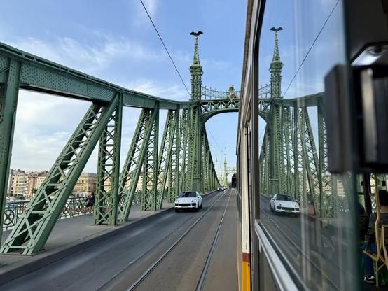 Blick aus dem Tramfenster bei der Fahrt über die Szabadság hid. Ein Teil der Brüxke und das entgegenkommende Auto spiegeln sich im Fenster der Tram.