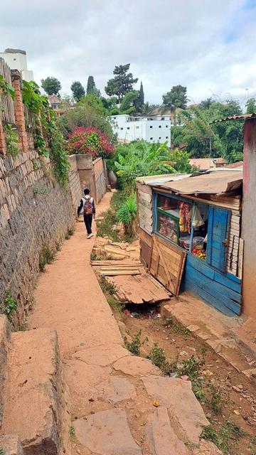 A narrow dirt path winds between a stone wall and a small wooden shop with a blue window shutter in a residential neighborhood in Madagascar. A person with a backpack walks away down the path, and the shop displays snacks, tomatoes, and other goods. Greenery and simple homes surround the area